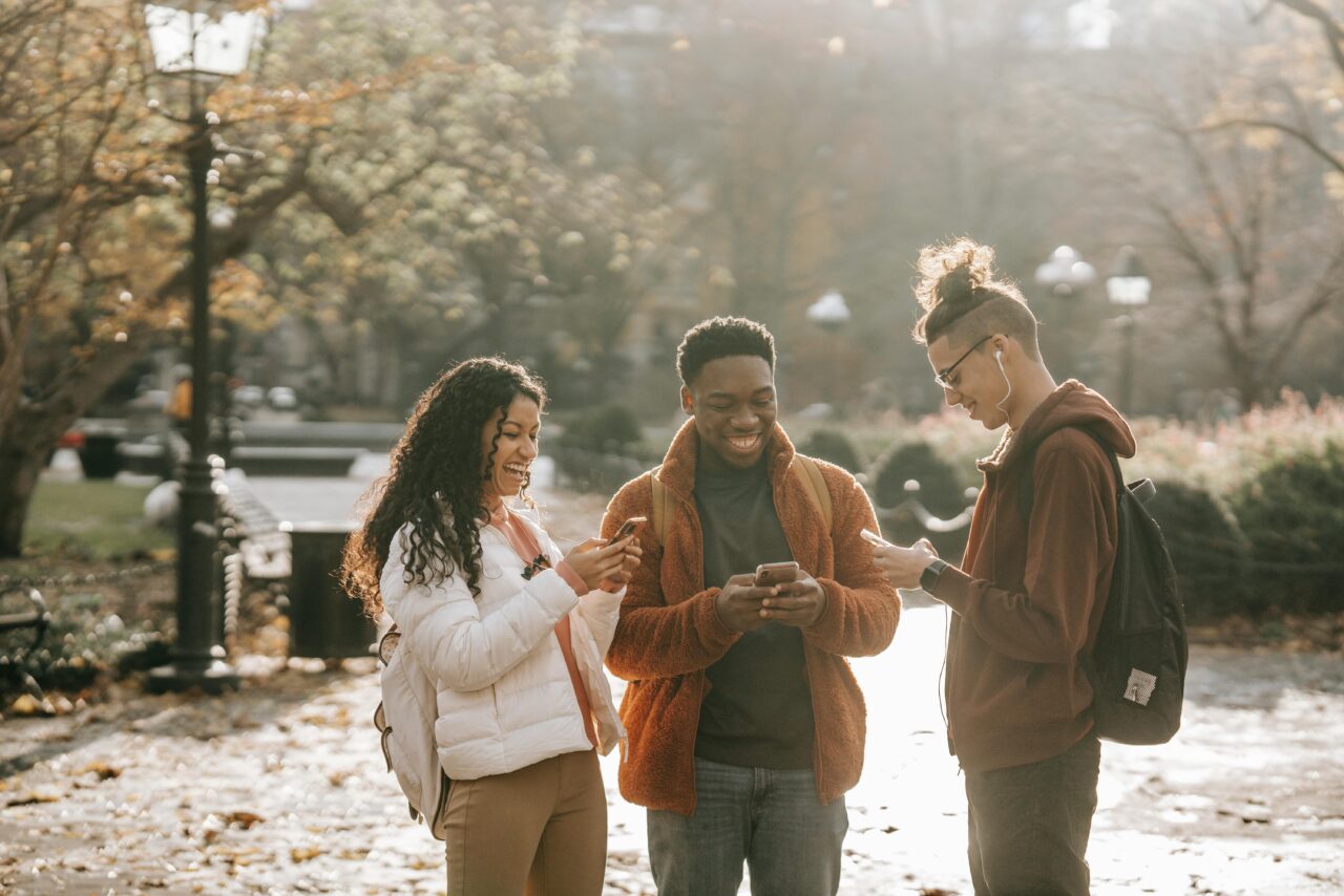 Three students standing together outdoors, smiling as they look at their phones